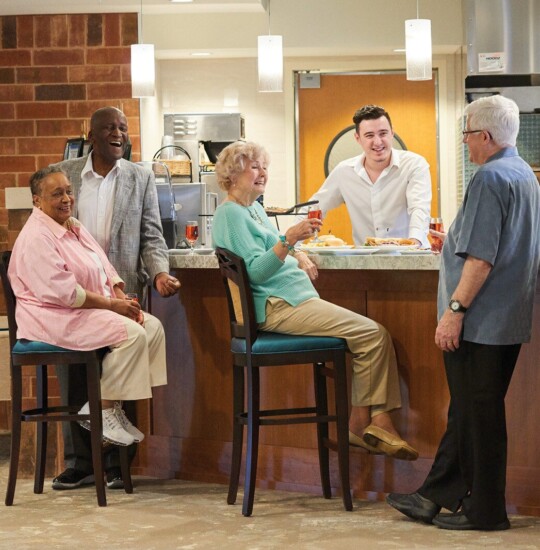 group of senior friends sit at the kitchen bar of the community dining area and converse