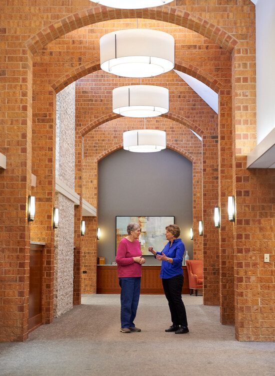 two senior women converse while standing in a hallway with beautiful arches at Beacon Hill Senior Living Community