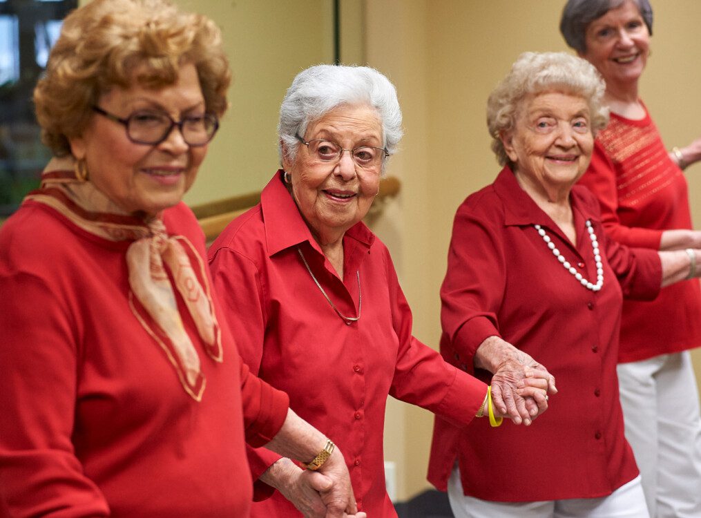 group of senior women in red shirts smile and hold hands while standing in a line