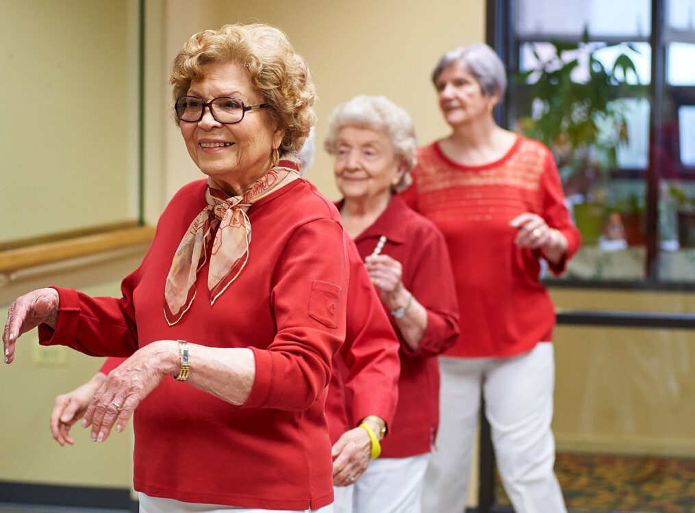 group of three senior women in red shirts stand next to a ballet bar during a dance class at Beacon Hill Senior Living Community