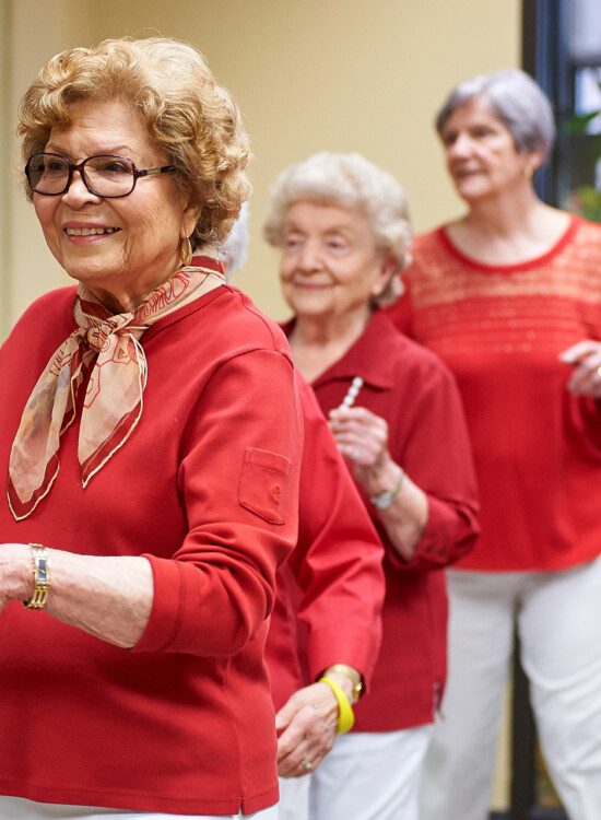 group of three senior women in red shirts stand next to a ballet bar during a dance class at Beacon Hill Senior Living Community