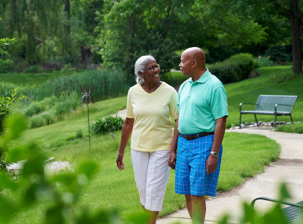 senior couple in casual summer clothes smile while walking on a scenic walking path surrounded by green landscaping