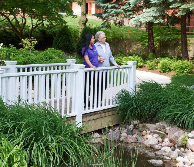 senior woman and her care giver stand on small bridge over the creek at Beacon Hill Senior Living Community, smiling at the lush green view