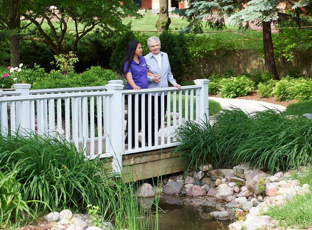 senior woman and her care giver stand on small bridge over the creek at Beacon Hill Senior Living Community, smiling at the lush green view