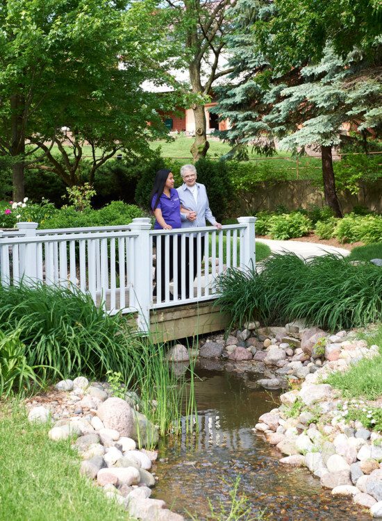 senior woman and her care giver stand on small bridge over the creek at Beacon Hill Senior Living Community, smiling at the lush green view