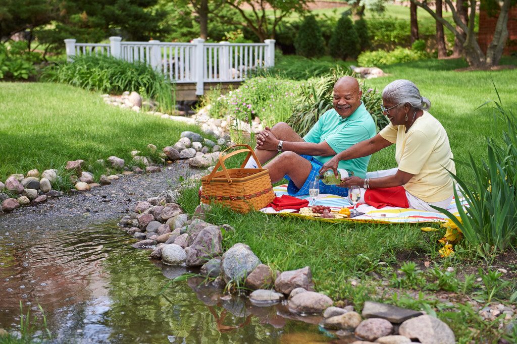 senior couple smile and enjoy an outdoor picnic in the grass beside a small creek at Beacon Hill Senior Living Community