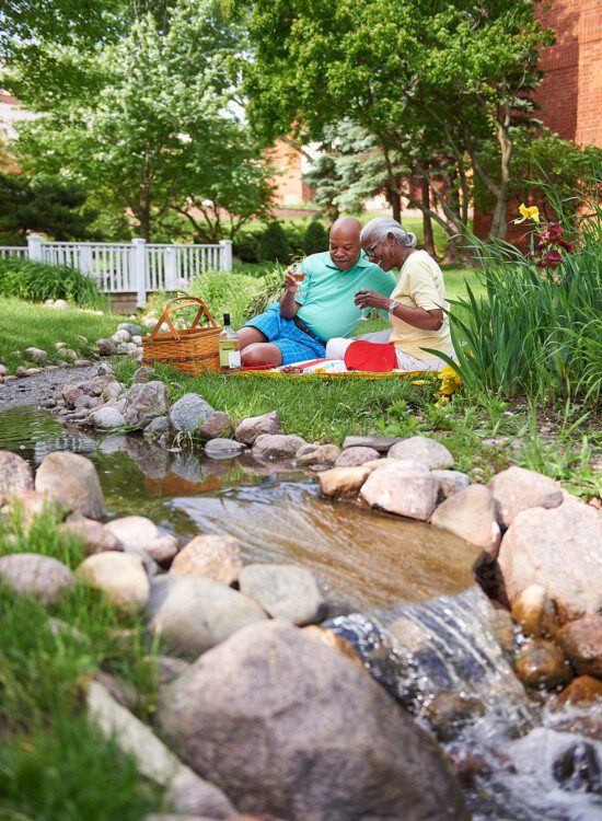 senior couple smile and enjoy an outdoor picnic in the grass beside a small creek at Beacon Hill Senior Living Community