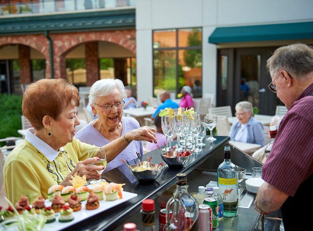 two senior women smile while enjoying cocktails and appetizers at the outdoor lounge and bar at Beacon Hill