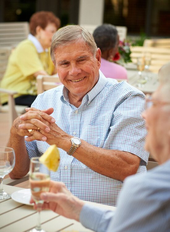 senior man smiles at his wife while dining outdoors at their senior living community