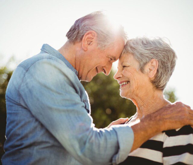 senior man and woman smile at each other while touching foreheads in an embrace outdoors