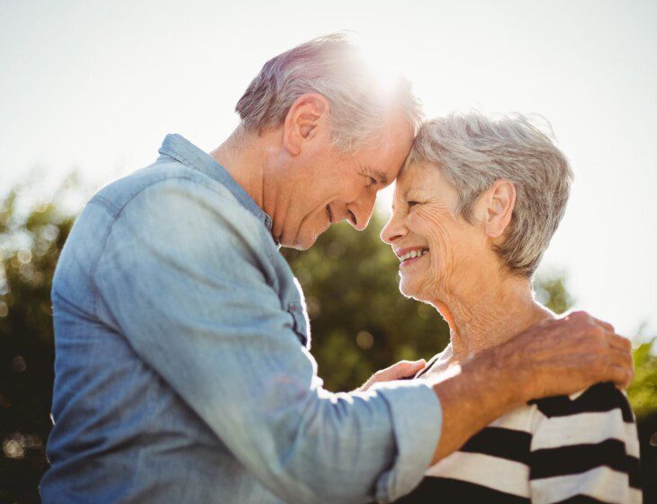 senior man and woman smile at each other while touching foreheads in an embrace outdoors