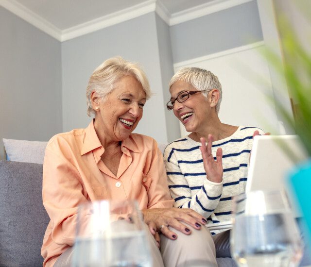 two senior women laugh and converse while seated on a couch indoors