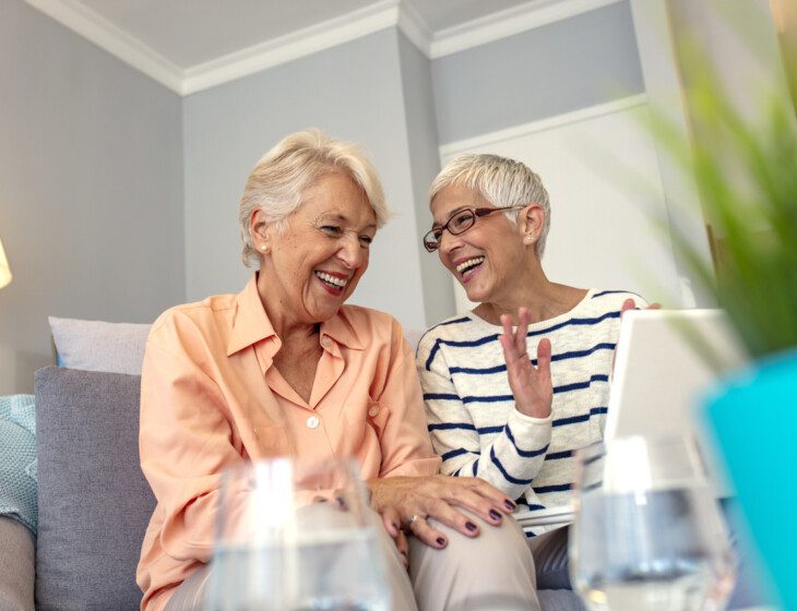 two senior women laugh and converse while seated on a couch indoors