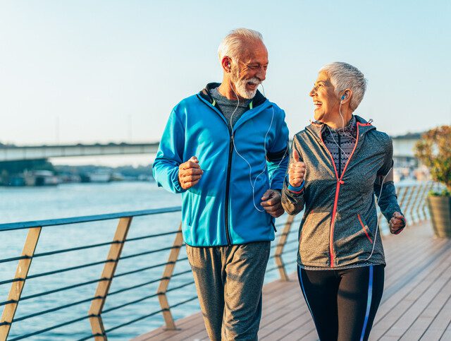 senior couple wearing athletic jackets and headphones smile while on a power walk together along a riverfront