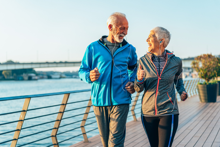 senior couple wearing athletic jackets and headphones smile while on a power walk together along a riverfront