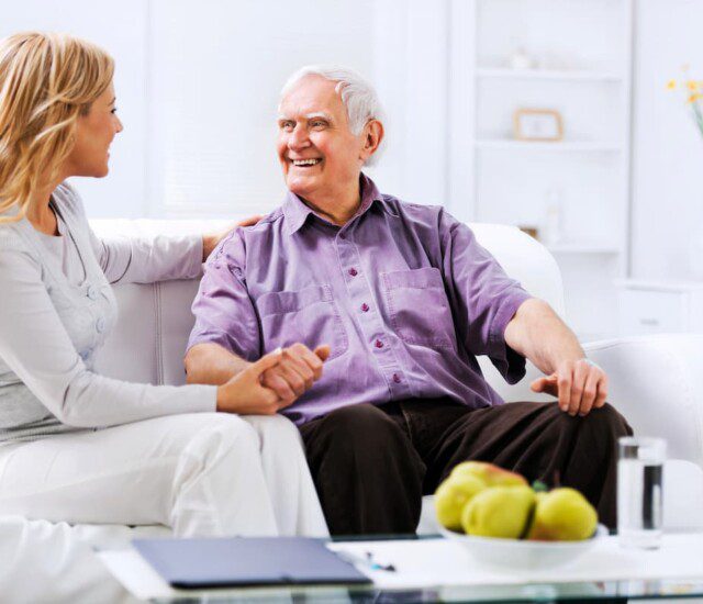 seated senior man on couch smiles and holds the hand of his adult daughter while they converse