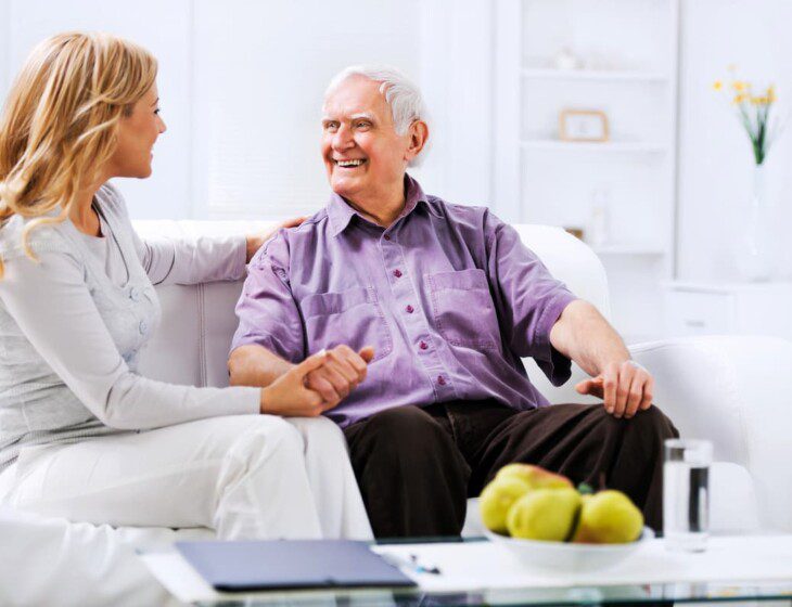 seated senior man on couch smiles and holds the hand of his adult daughter while they converse