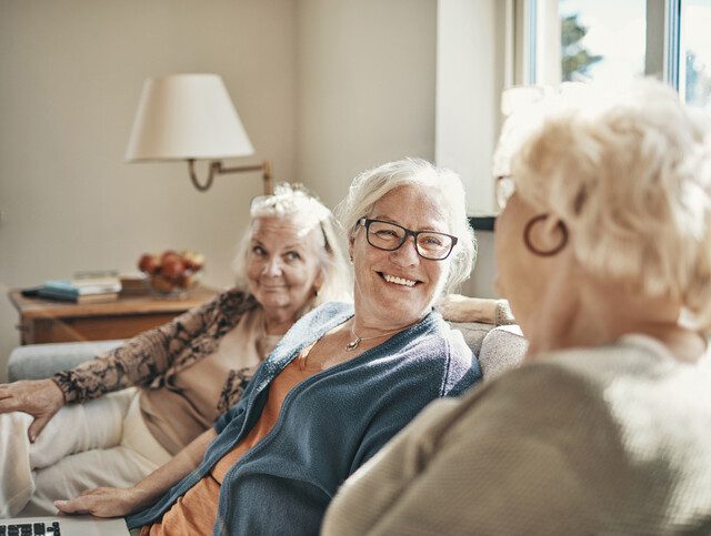 group of three senior women smile and converse while seated on a couch indoors