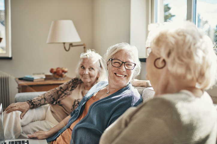 group of three senior women smile and converse while seated on a couch indoors