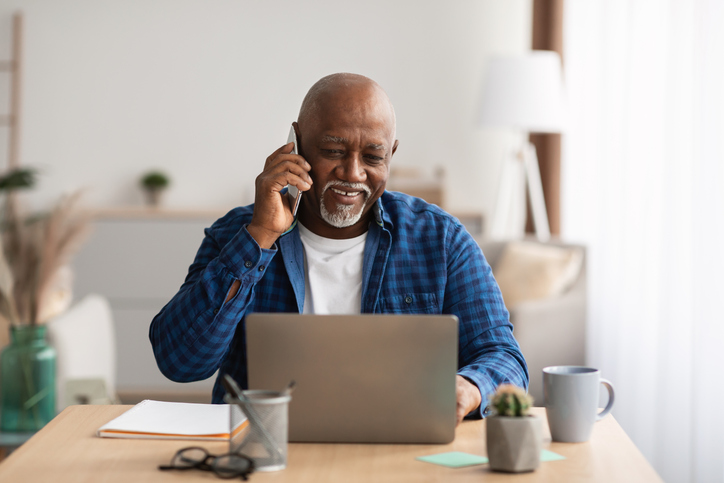 senior man sitting at his table on the phone, looking at his laptop and smiling