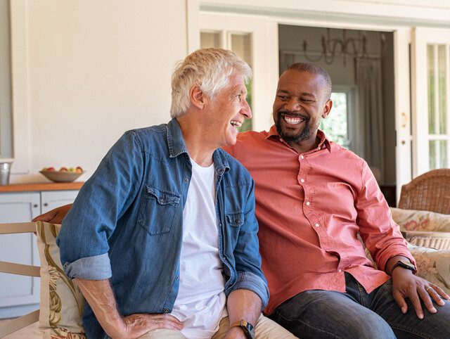 two senior men smile and sit on a couch together