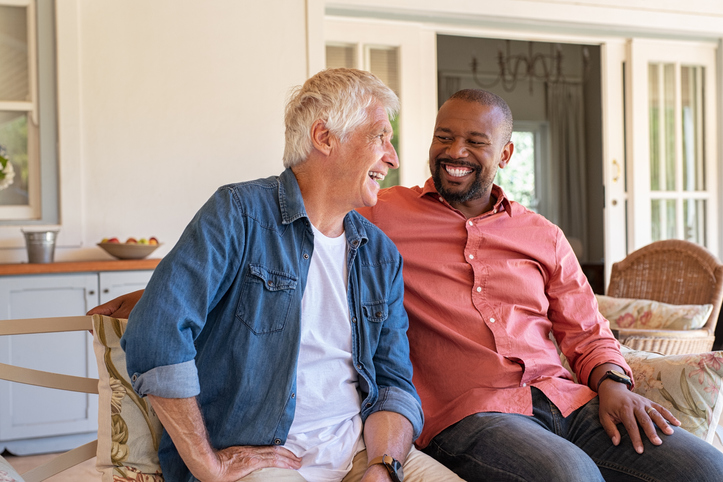 two senior men smile and sit on a couch together