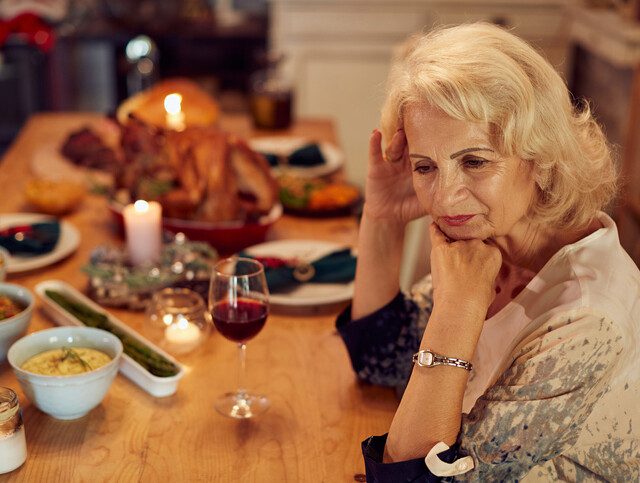 senior woman sits alone at a table set for Thanksgiving, looking forlorn
