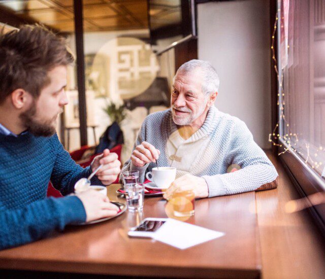 senior man and his adult son smile while conversing over coffee at a local cafe