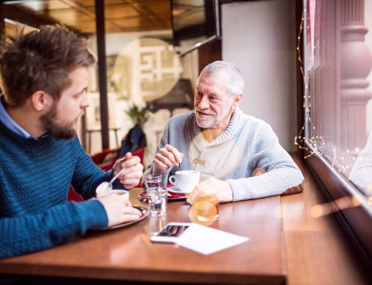 senior man and his adult son smile while conversing over coffee at a local cafe