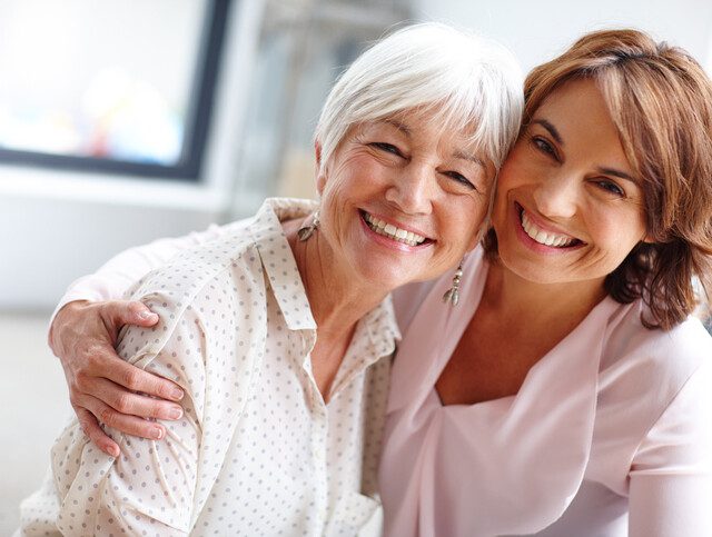 senior woman and her adult daughter smile and look at the camera, their arms around one another