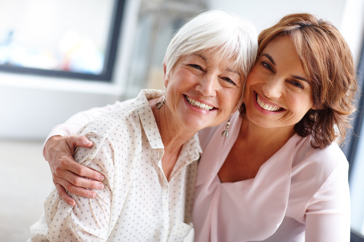 senior woman and her adult daughter smile and look at the camera, their arms around one another