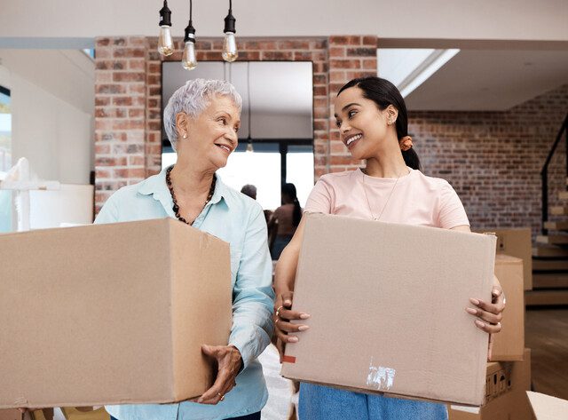 senior woman and her adult daughter each hold a medium moving box, looking at one another and smiling