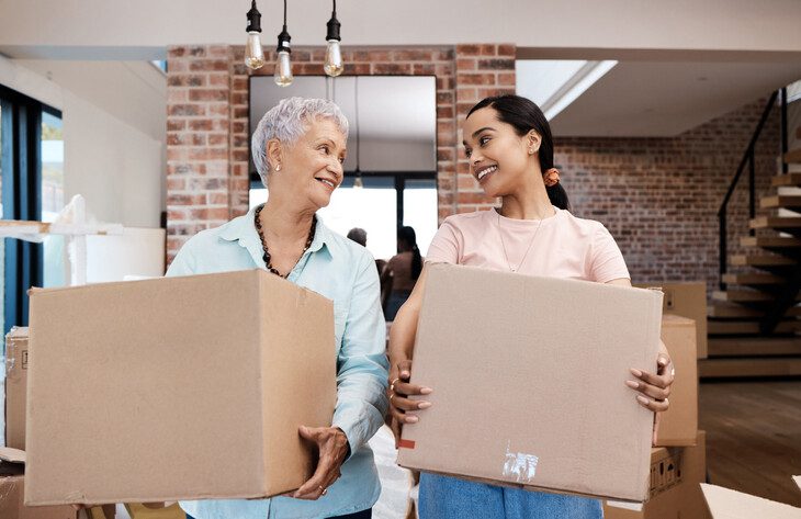 senior woman and her adult daughter each hold a medium moving box, looking at one another and smiling