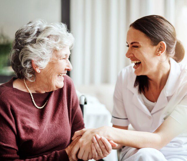 seated senior woman and her female caregiver laugh and hold hands while conversing