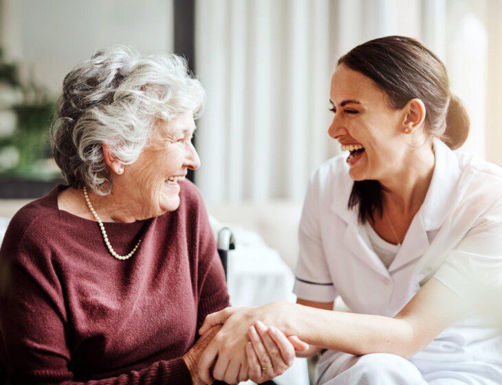 seated senior woman and her female caregiver laugh and hold hands while conversing